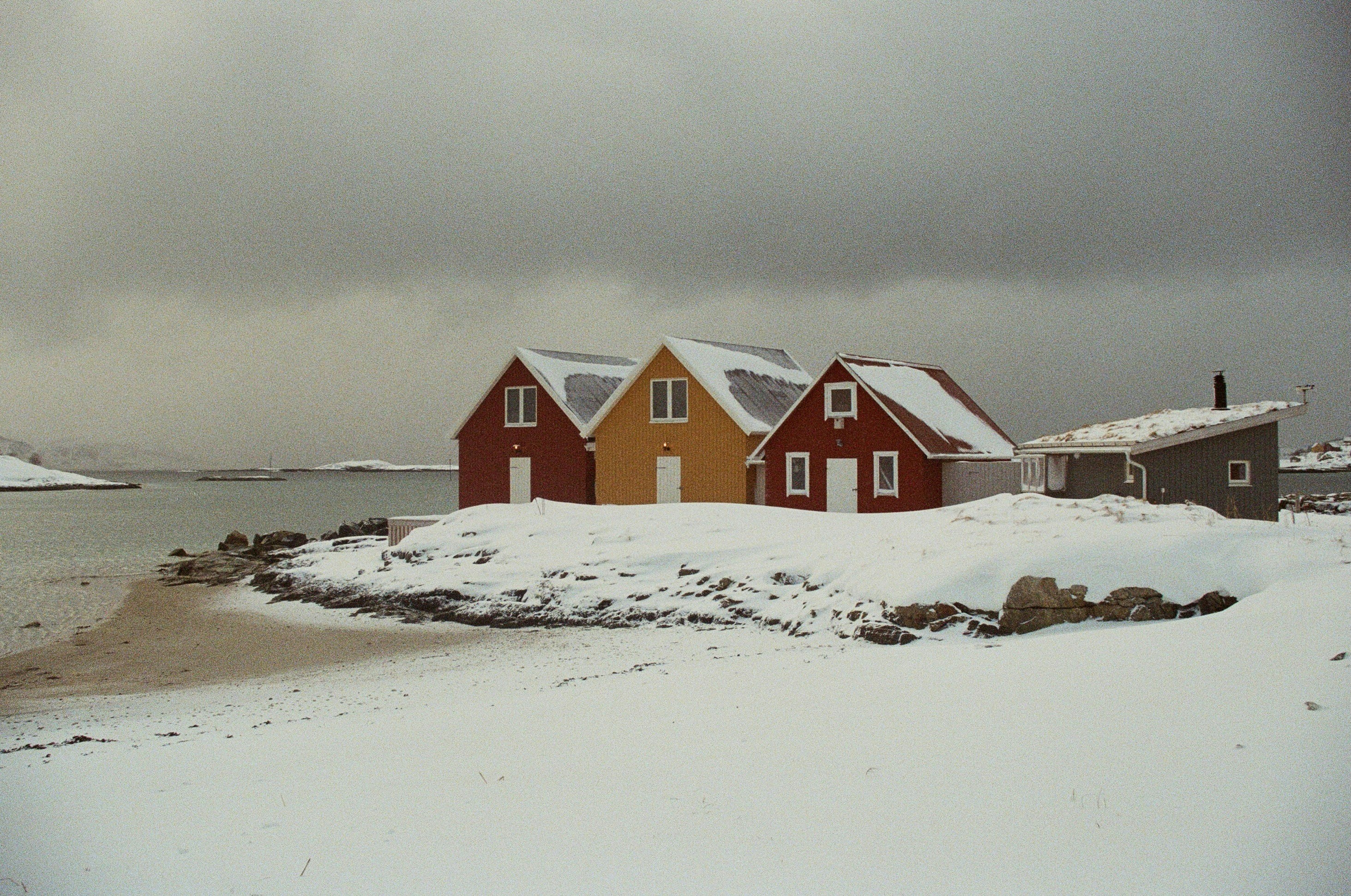 Three brightly painted cabins—red, yellow, and red again—sit on a snowy shoreline in Lofoten, Norway, beneath a moody gray sky