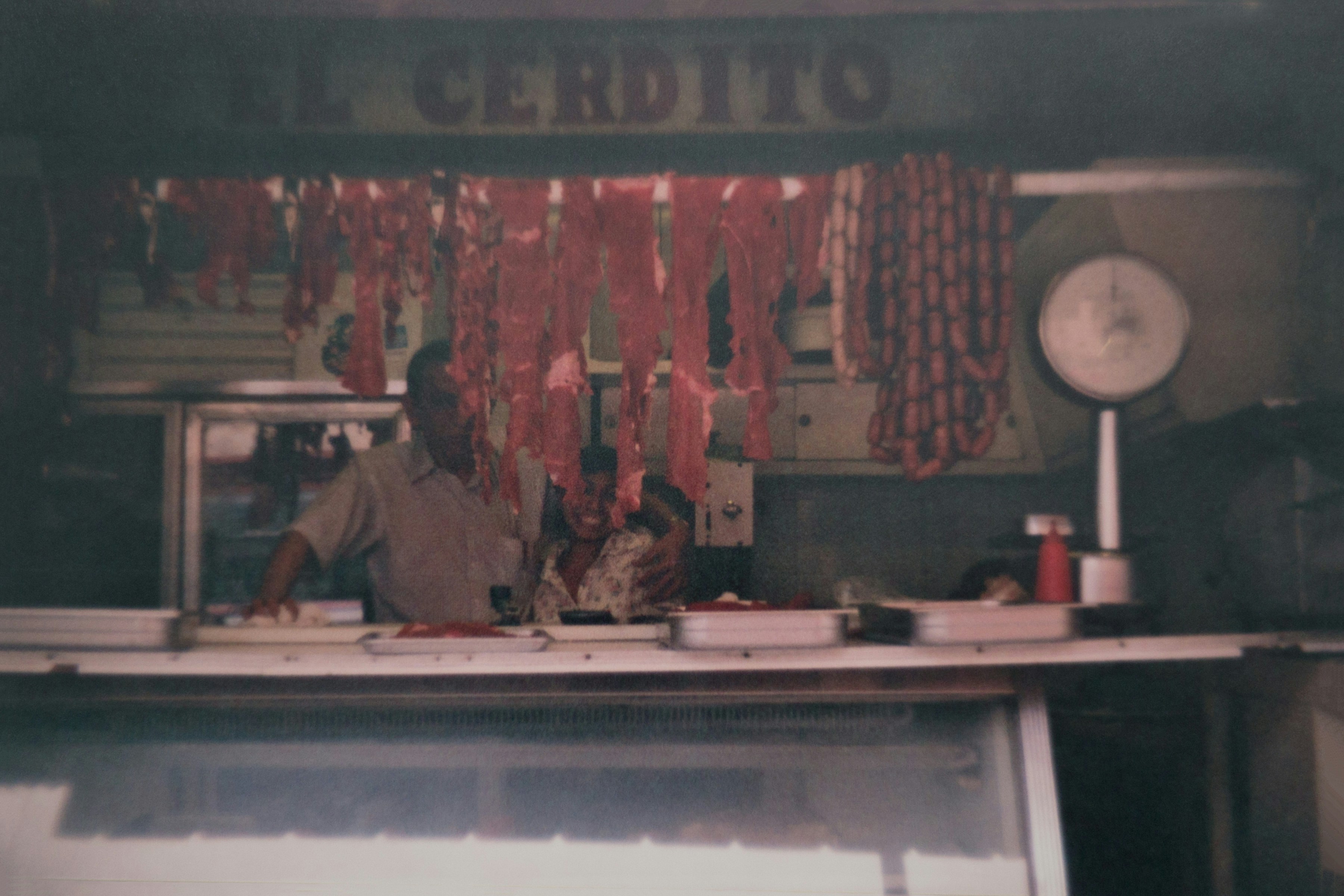 Dimly lit butcher shop named “El Cerdito,” with meat and sausages hanging from hooks, a scale on the counter, and two people smiling behind the display case.