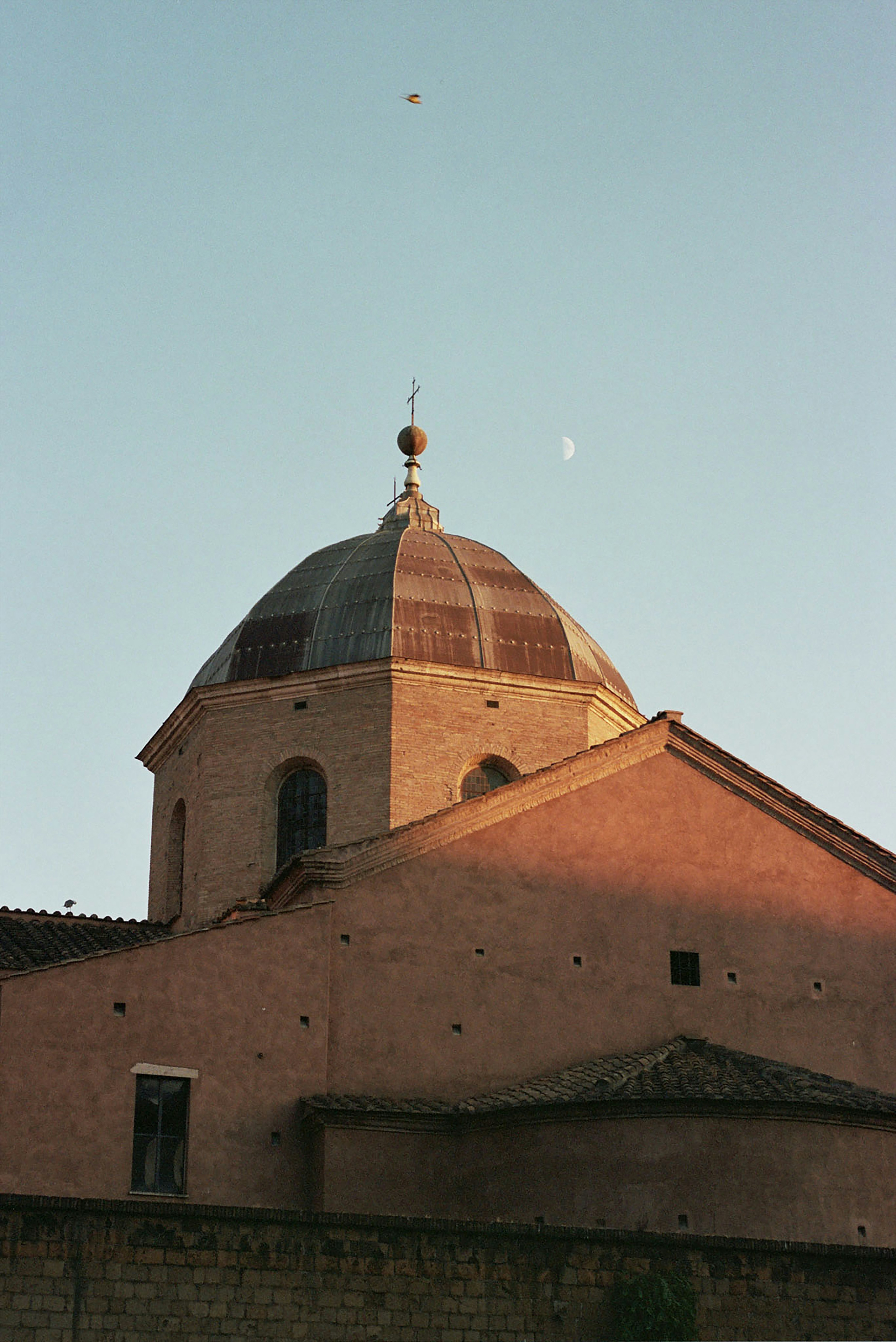 Sunlit dome of a historic church in Rome at dusk, with the moon visible in the clear evening sky above the terracotta-colored building.