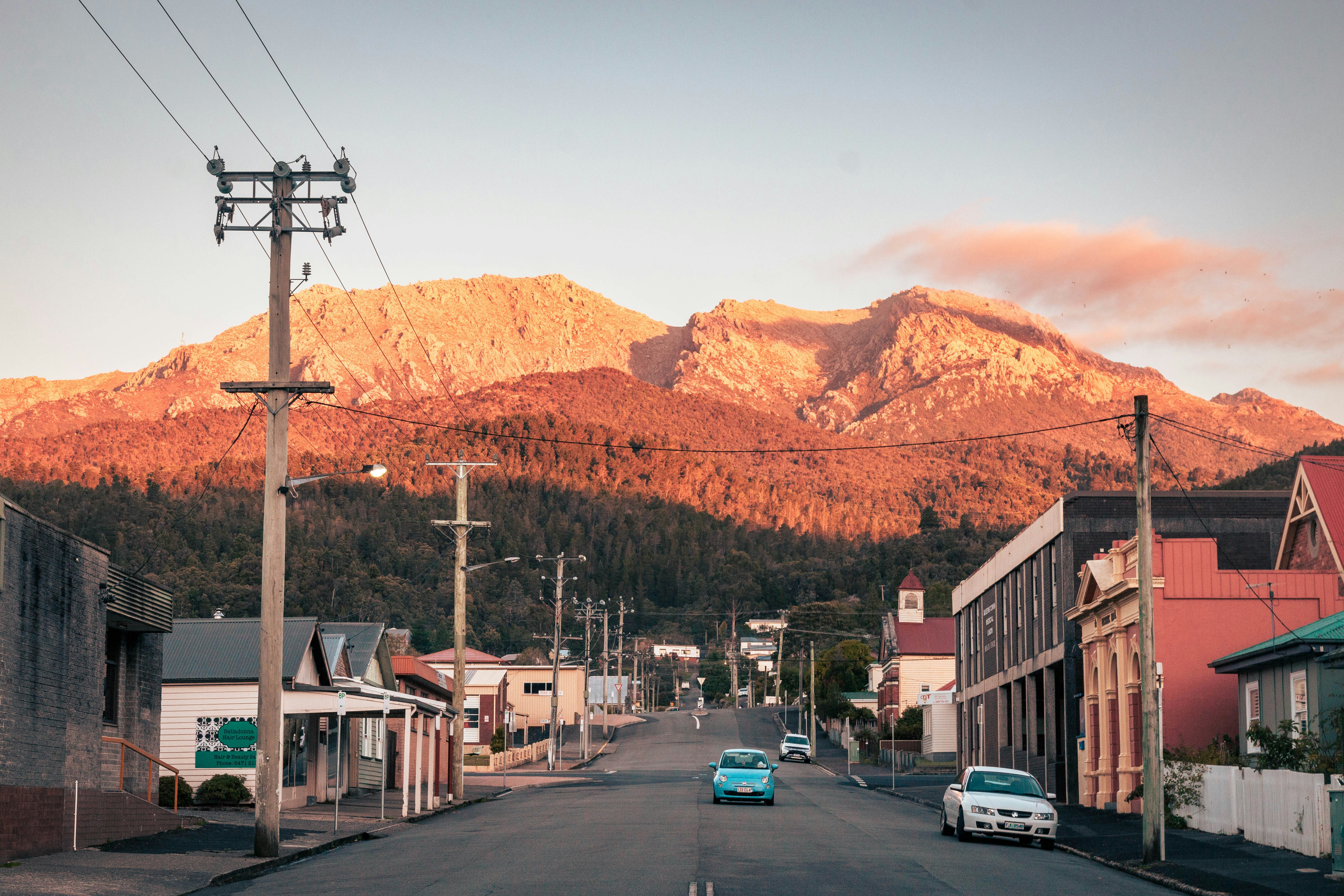 Quiet street in a Tasmanian town with cars parked along the road, set against a backdrop of rugged mountains glowing orange in the late afternoon sun.