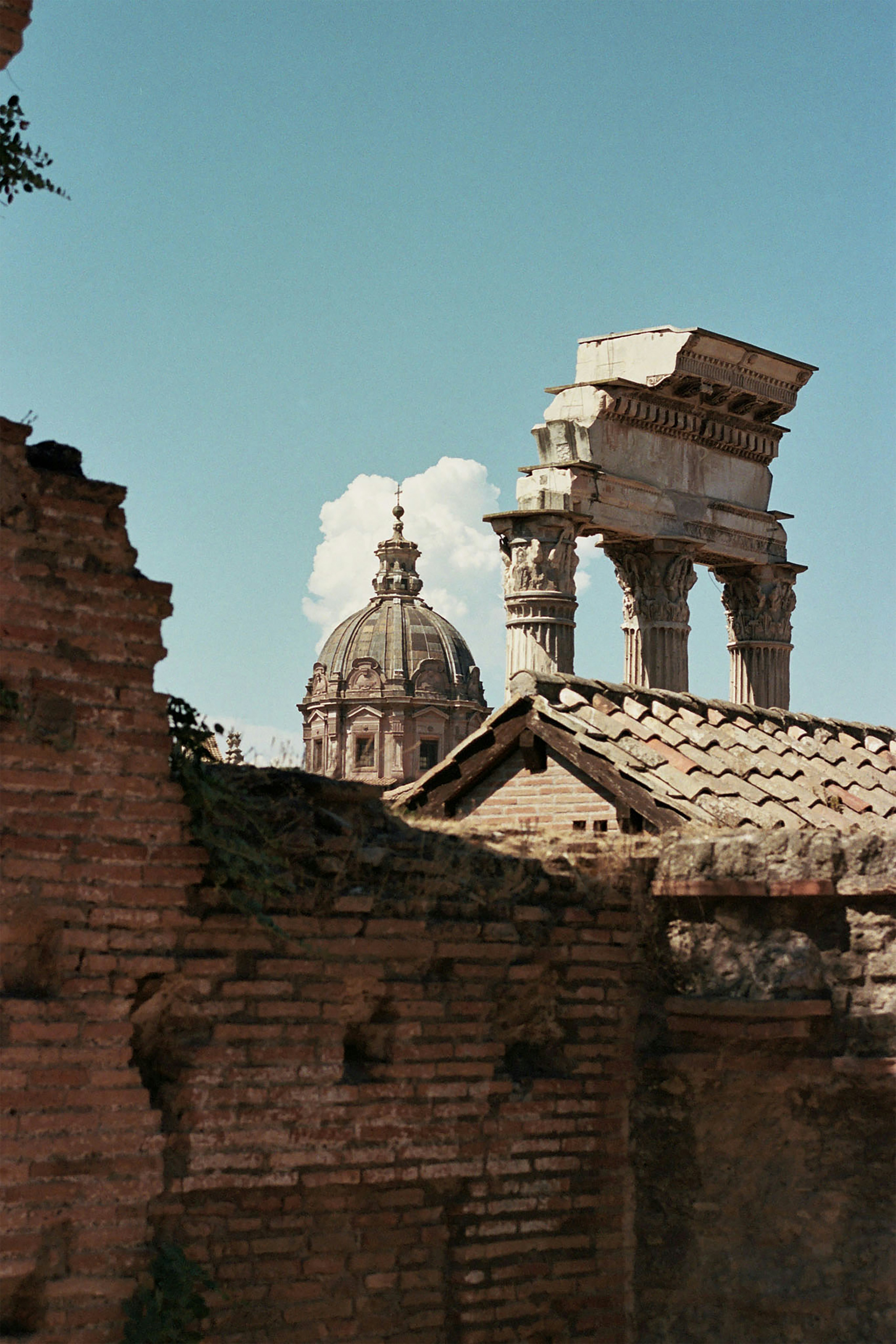 View of ancient Roman ruins with brick walls and stone columns in the foreground, framing the dome of a Baroque church under a bright blue sky.