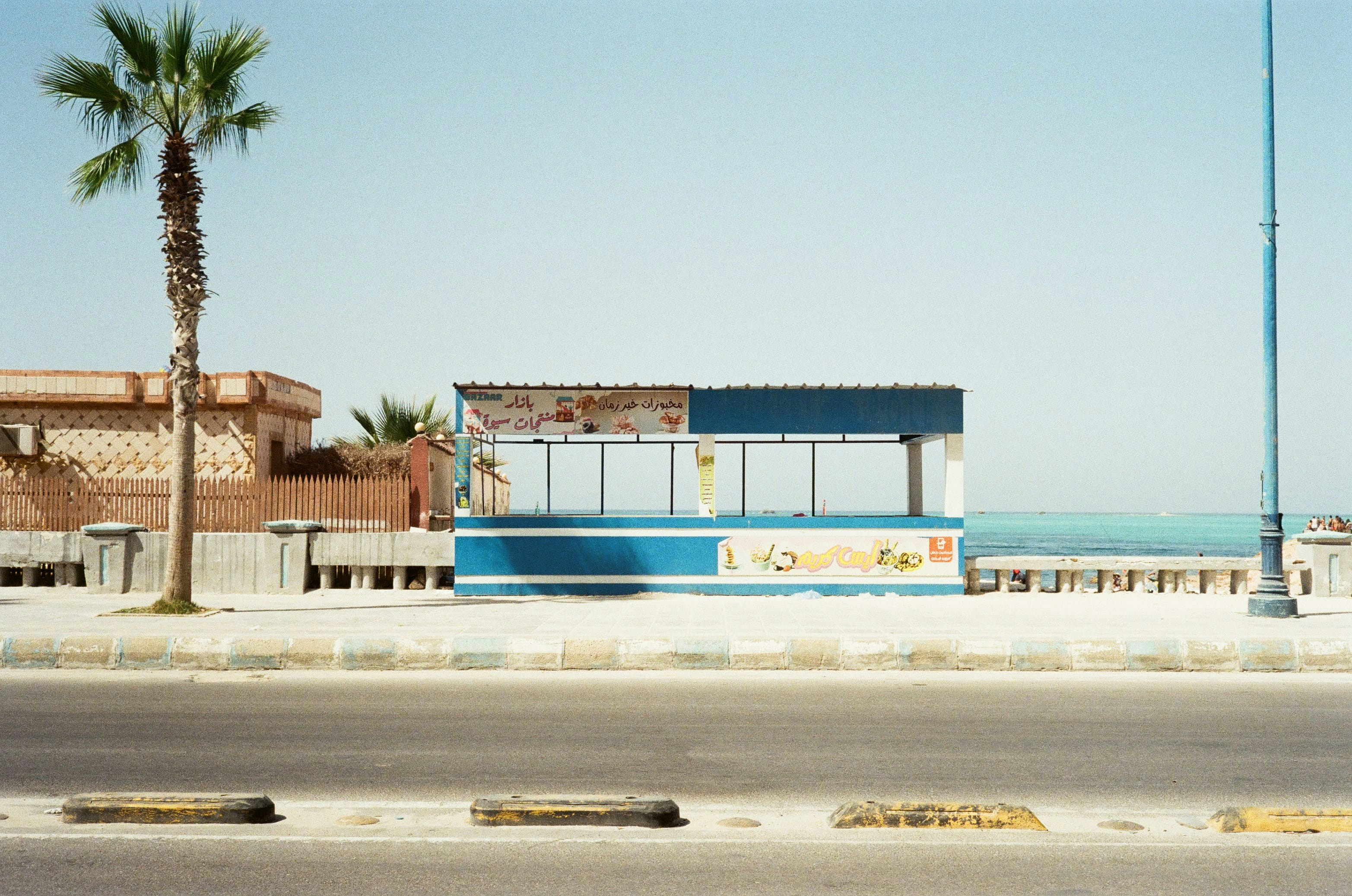 A quiet roadside snack stand painted blue and white sits empty by the shore of the Red Sea under a pale sky, framed by palm trees and a street lamp